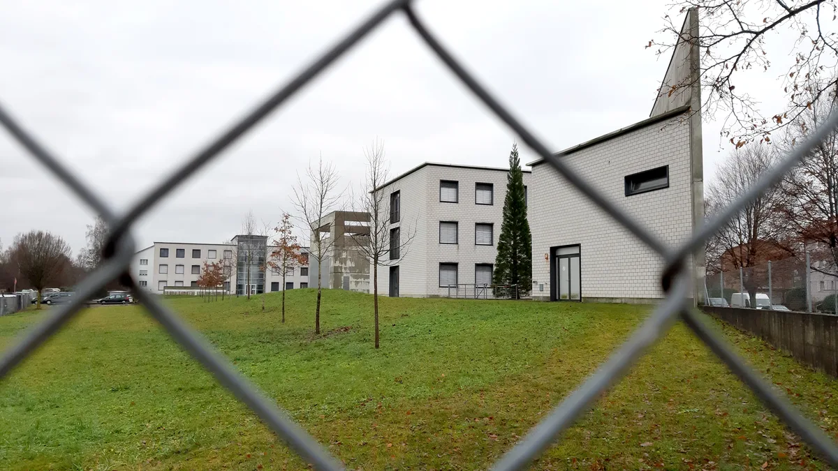 Ein mehrteiliges Gebäude hinter einem Maschendrahtzaun. 